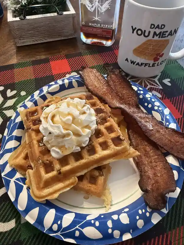 A plate of waffles topped with whipped cream and syrup, accompanied by crispy bacon, sits on a blue and white patterned…