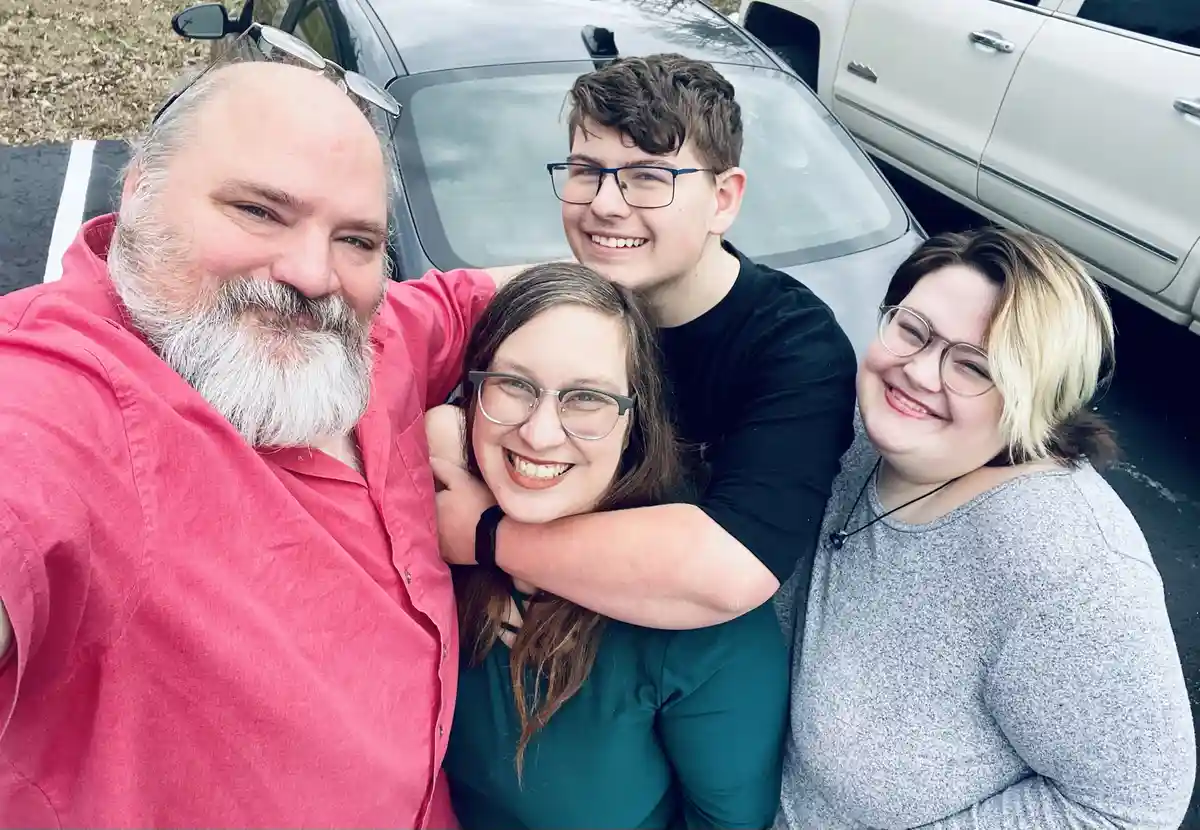Four people, including a man with a white beard in a red shirt, are smiling and hugging in front of a car. The overlaid text…