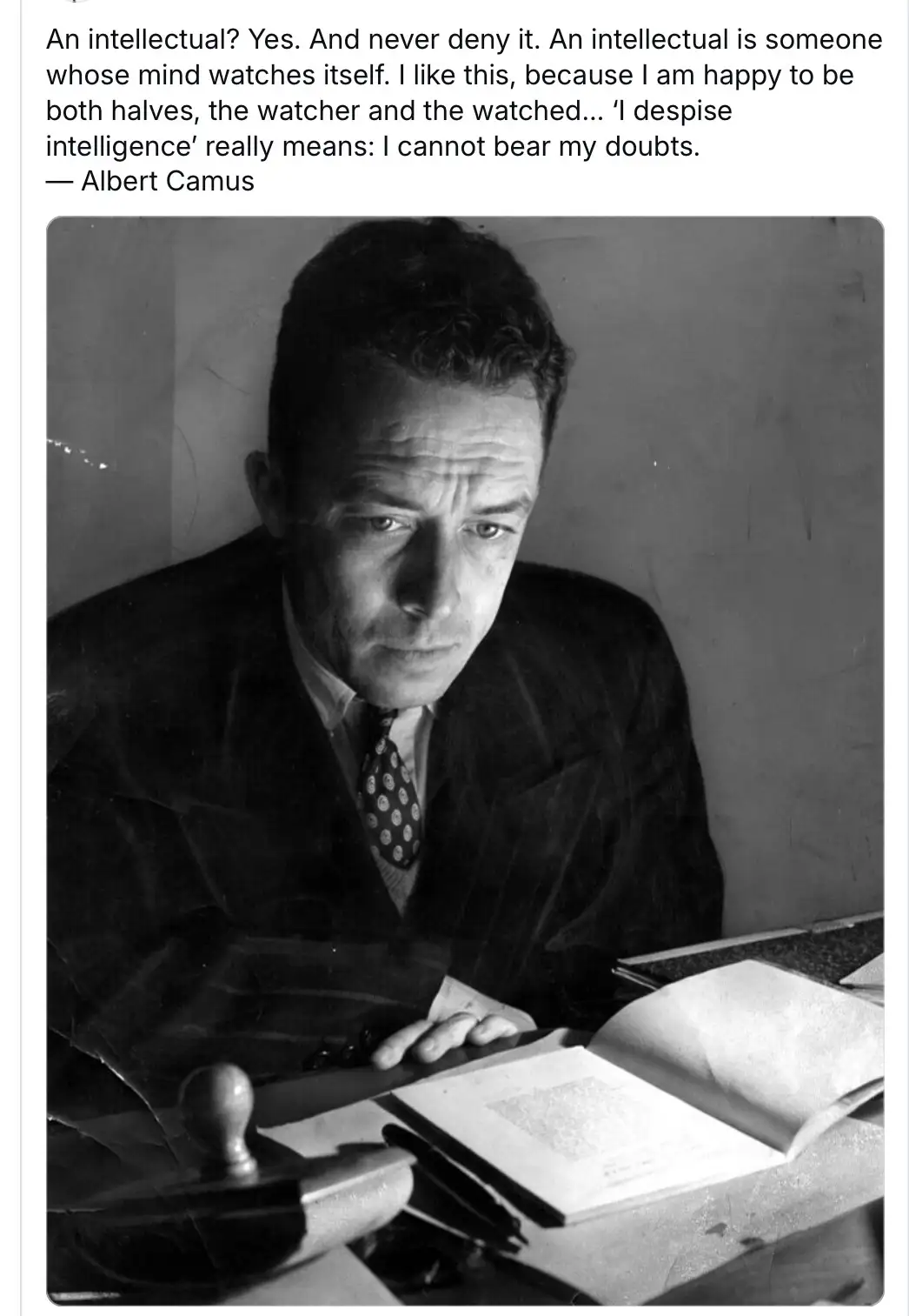 A black-and-white photograph shows a man in a suit and tie sitting at a desk with an open book, looking intently at the…