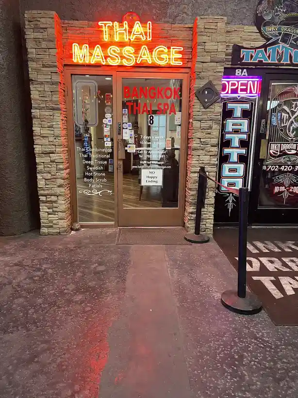 A storefront with a bright red neon sign reading 'THAI MASSAGE' above glass doors for 'BANGKOK THAI SPA'. A small white sign…