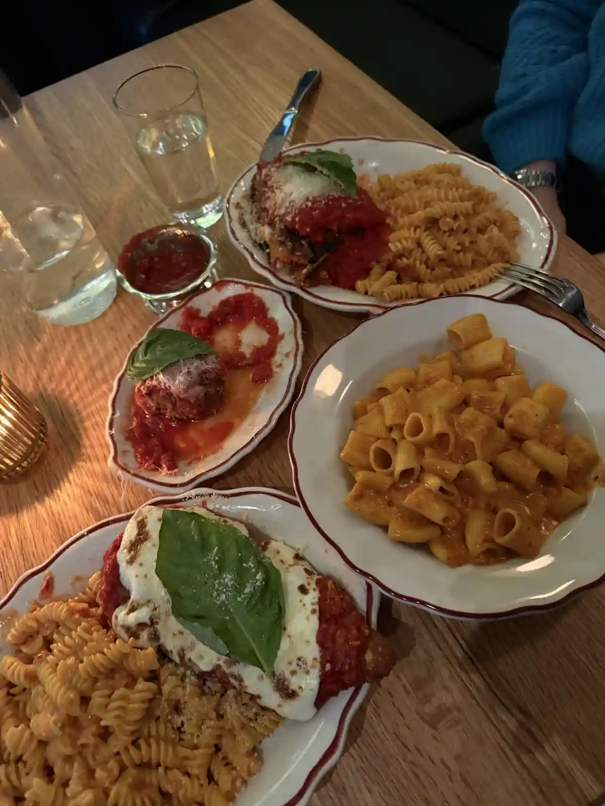 A wooden table is covered with four plates of Italian food: two servings of chicken parmesan with pasta, a plate of rigatoni…