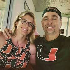 A smiling man and woman pose for a selfie indoors. The man wears a black t-shirt with a large orange 'U' logo and a black…