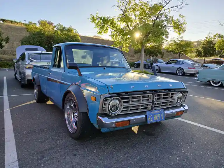 A light blue vintage Ford pickup truck with chrome wheels and a California license plate is parked in a lot surrounded by…