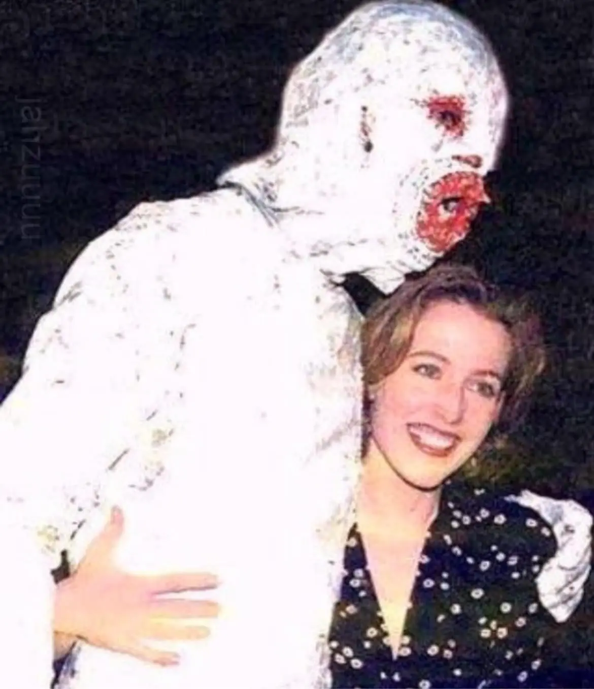 A woman in a black patterned top smiles brightly while being embraced from behind by a person in a full-body white costume…