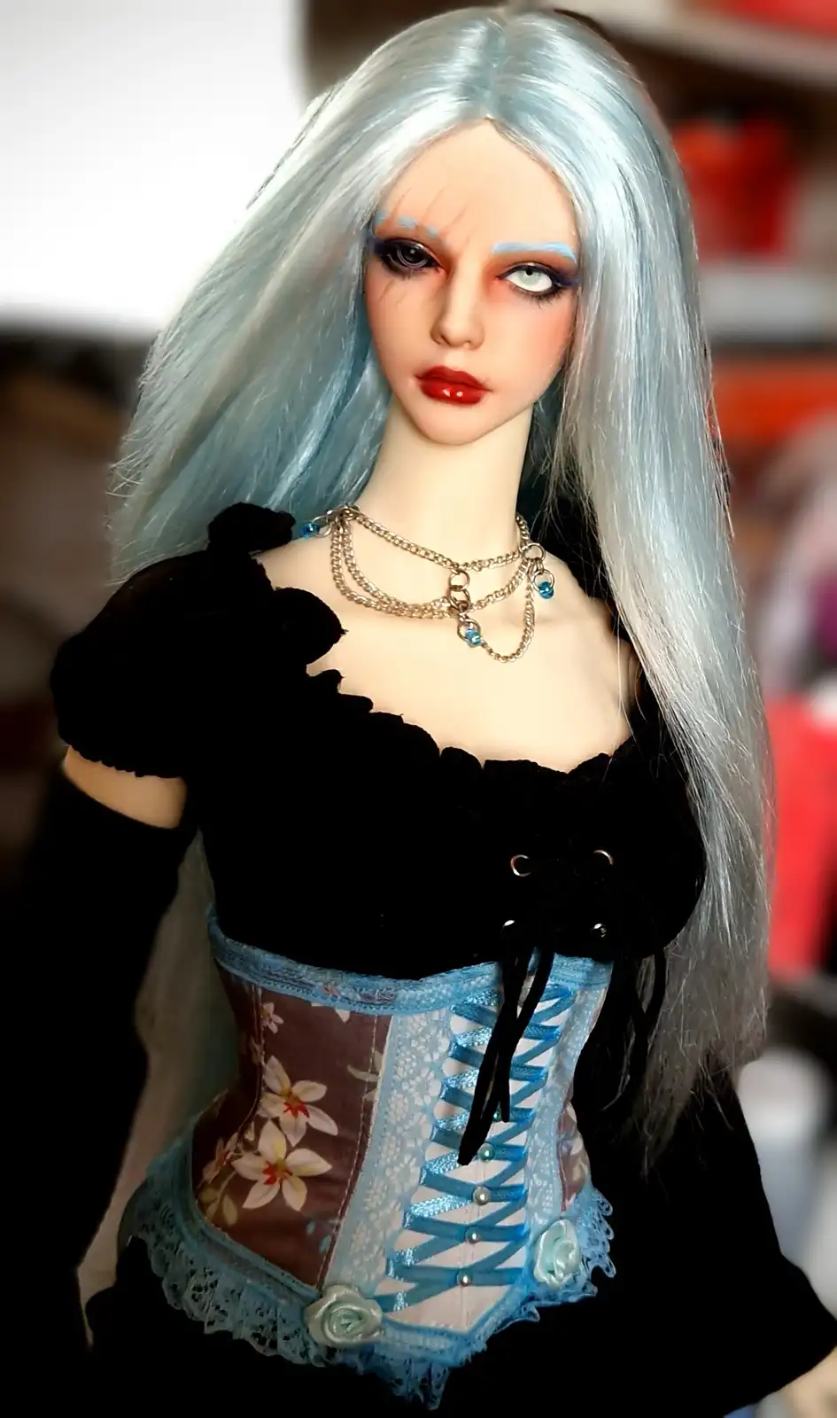 A close-up of a doll with long, straight, light blue hair, dramatic makeup including dark eyeliner and red lipstick, and a…