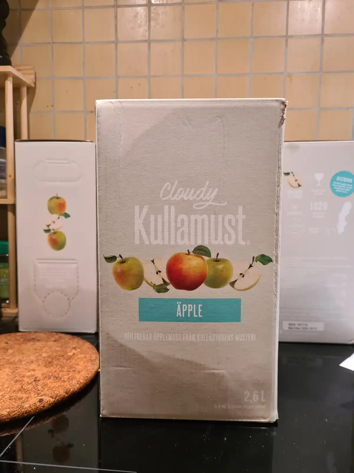 A large cardboard box of 'Cloudy Kullamust' apple must (unfiltered apple cider) sits on a black kitchen counter, with…