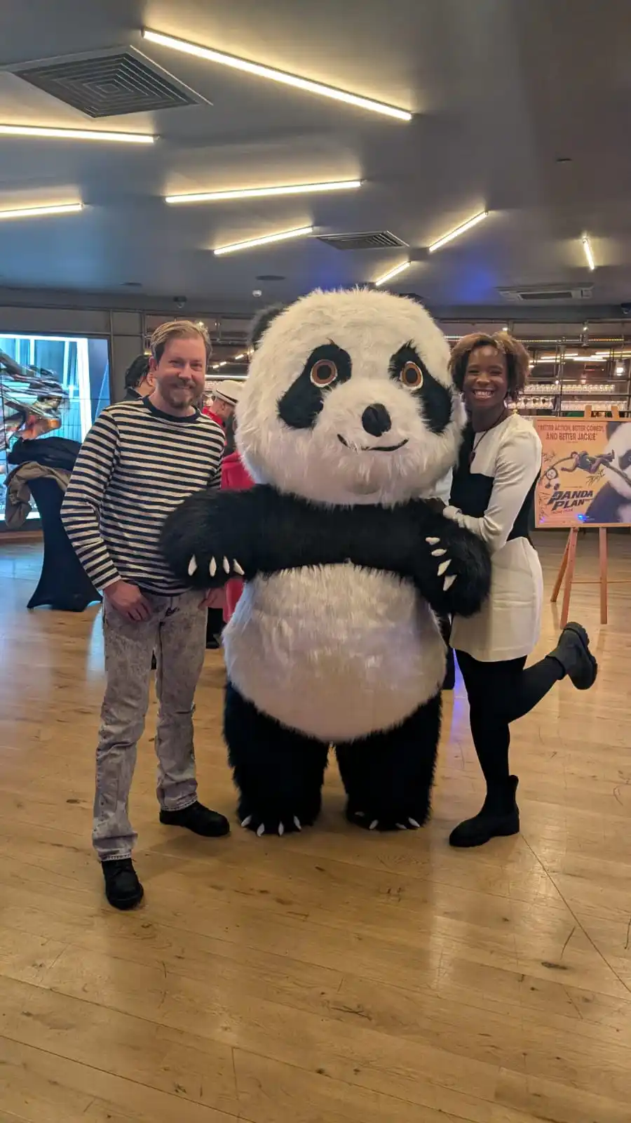 Two people pose with a large, fluffy panda mascot costume in a brightly lit indoor space with wooden floors. The man on the…