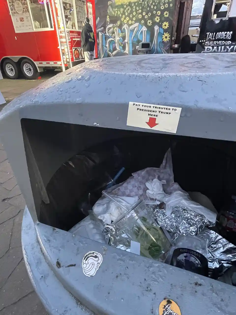 A close-up photo of a public trash can filled with discarded food wrappers and cups. A white sticker on the lid reads 'PAY…