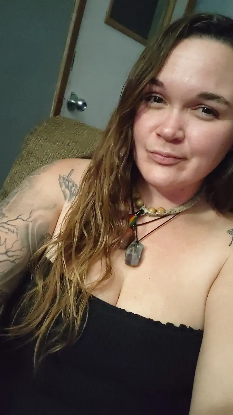 A selfie of a woman with long brown hair, wearing a black top and layered necklaces, showing tattoos on her arms. She has a…