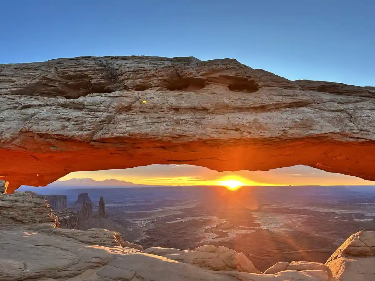 A stunning photograph captures a sunrise or sunset viewed through a large natural sandstone arch, with the sun glowing…