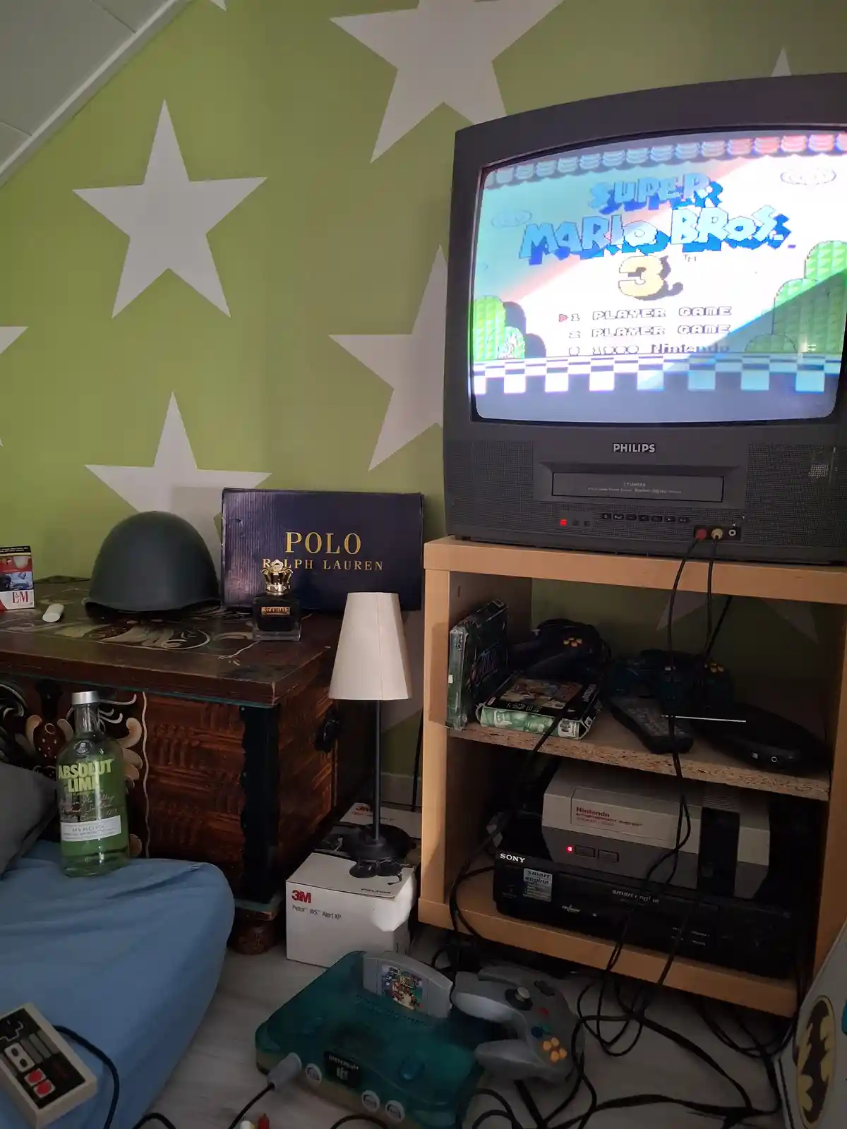 A cluttered room with a Philips CRT TV displaying the Super Mario Bros. 3 title screen, surrounded by vintage gaming…