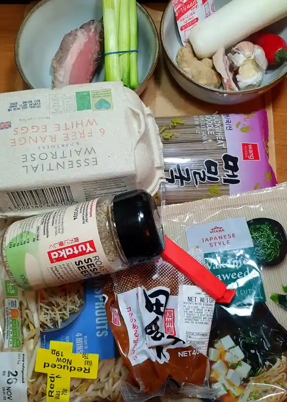 A flat-lay photo of various grocery items including a carton of eggs, buckwheat noodles, soy sauce, bean sprouts, seaweed…