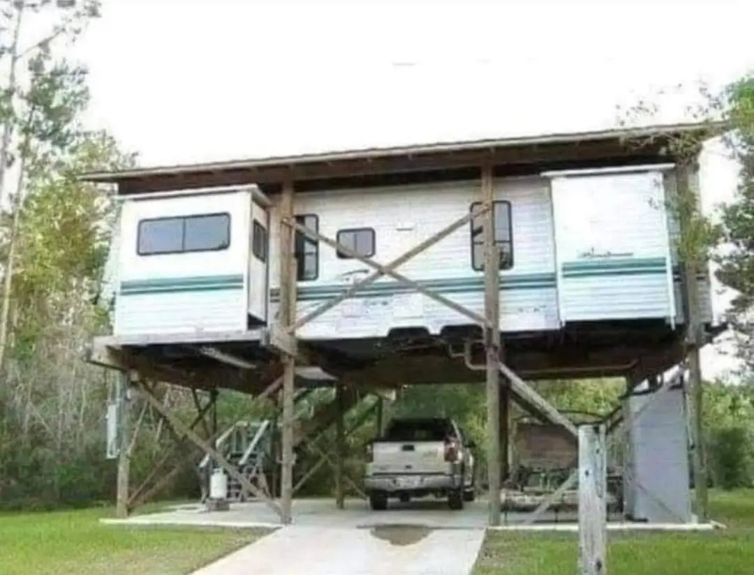 A white RV is elevated on a wooden platform with diagonal braces, supported by stilts, with a pickup truck parked…