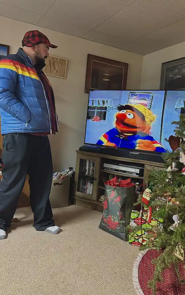 A man in a blue puffer jacket and red plaid hat stands in a living room, looking at a TV screen showing the Muppet character…