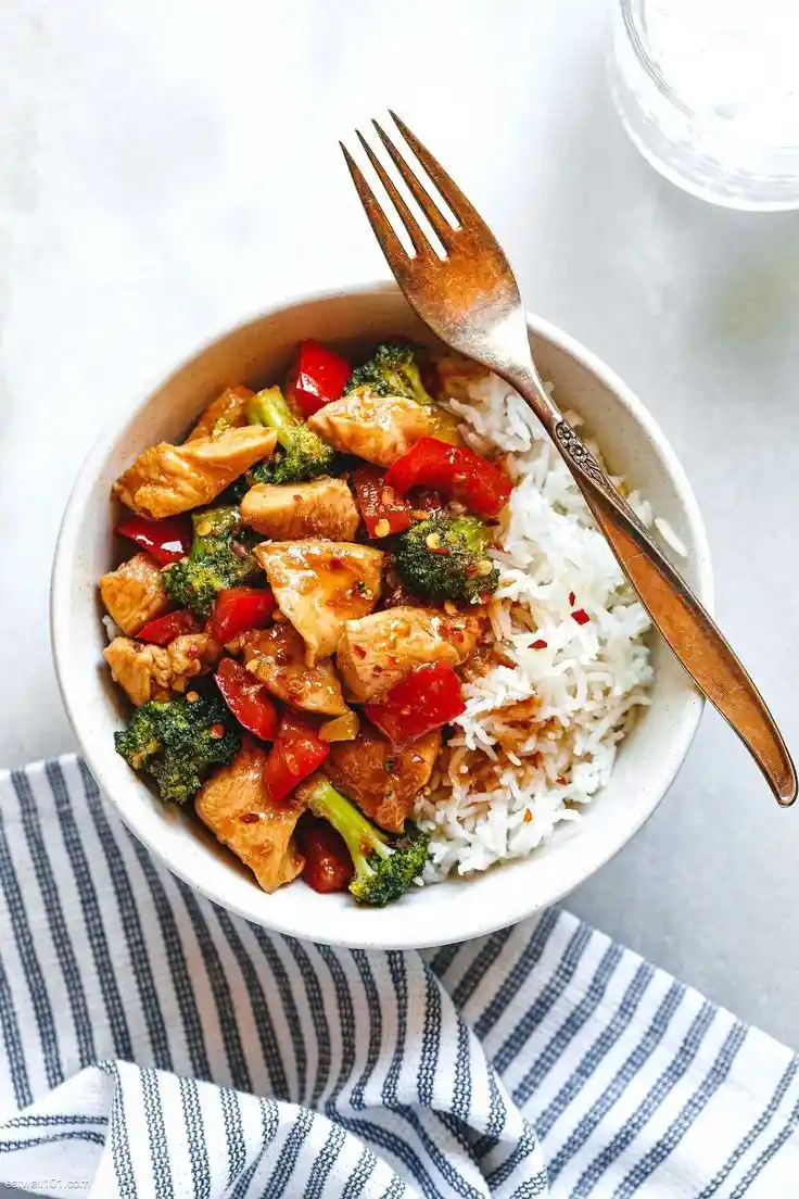 A top-down photo of a white bowl filled with chicken, broccoli, red bell peppers, and white rice, with a fork resting in the…