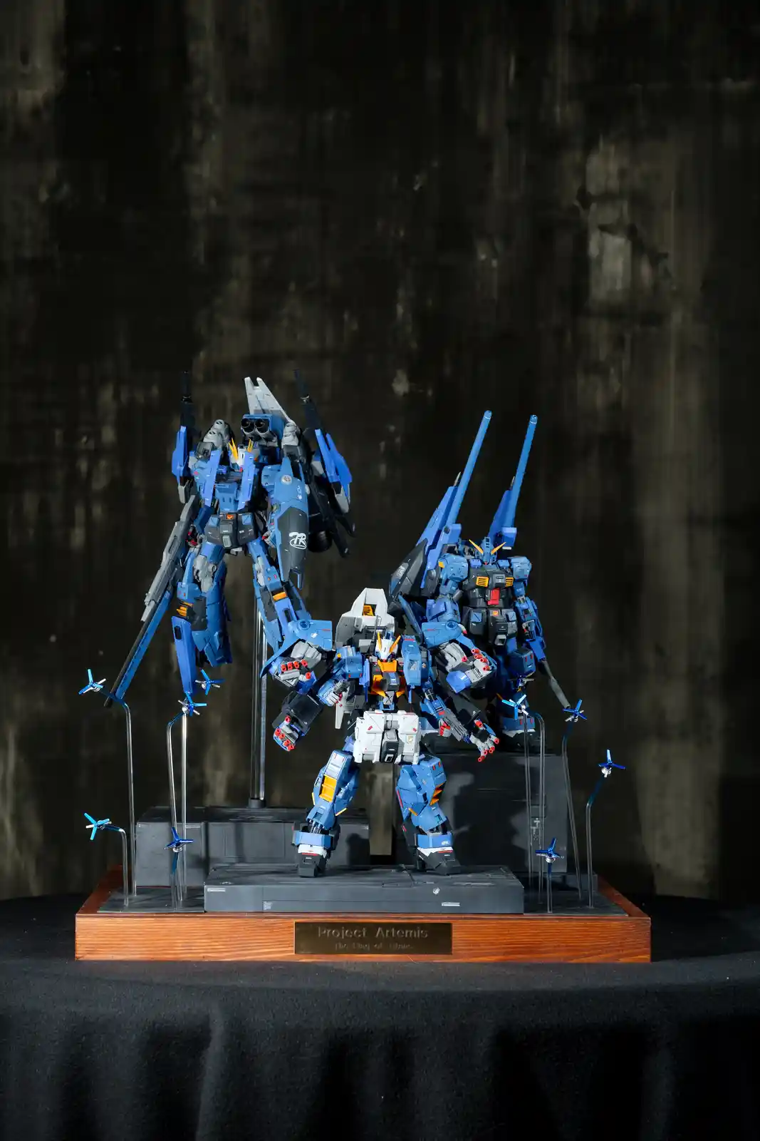 Three intricately detailed blue and white mecha model kits are displayed on a tiered black base with a wooden front panel…