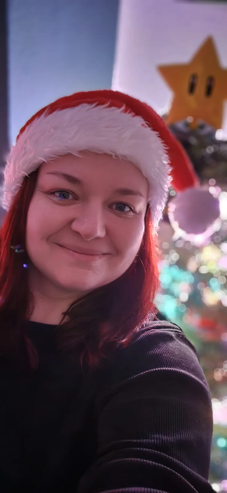 A person with red hair wearing a red and white Santa hat smiles at the camera, standing in front of a blurred Christmas tree…