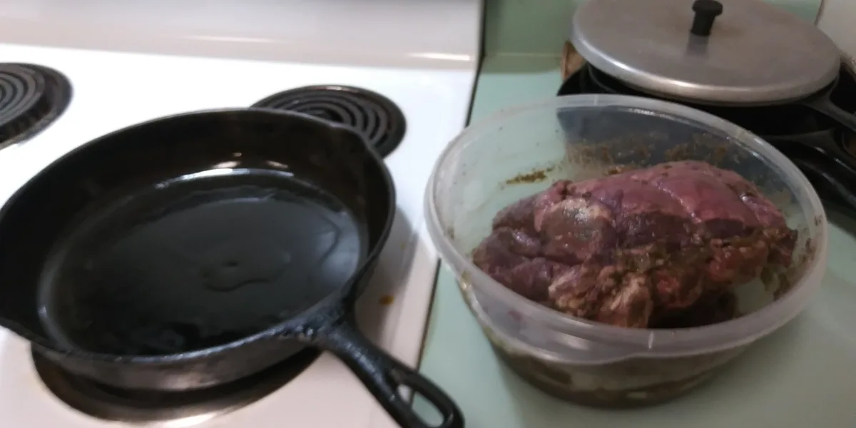 A black cast iron skillet sits on a white electric stovetop burner, next to a clear plastic container holding a large piece…