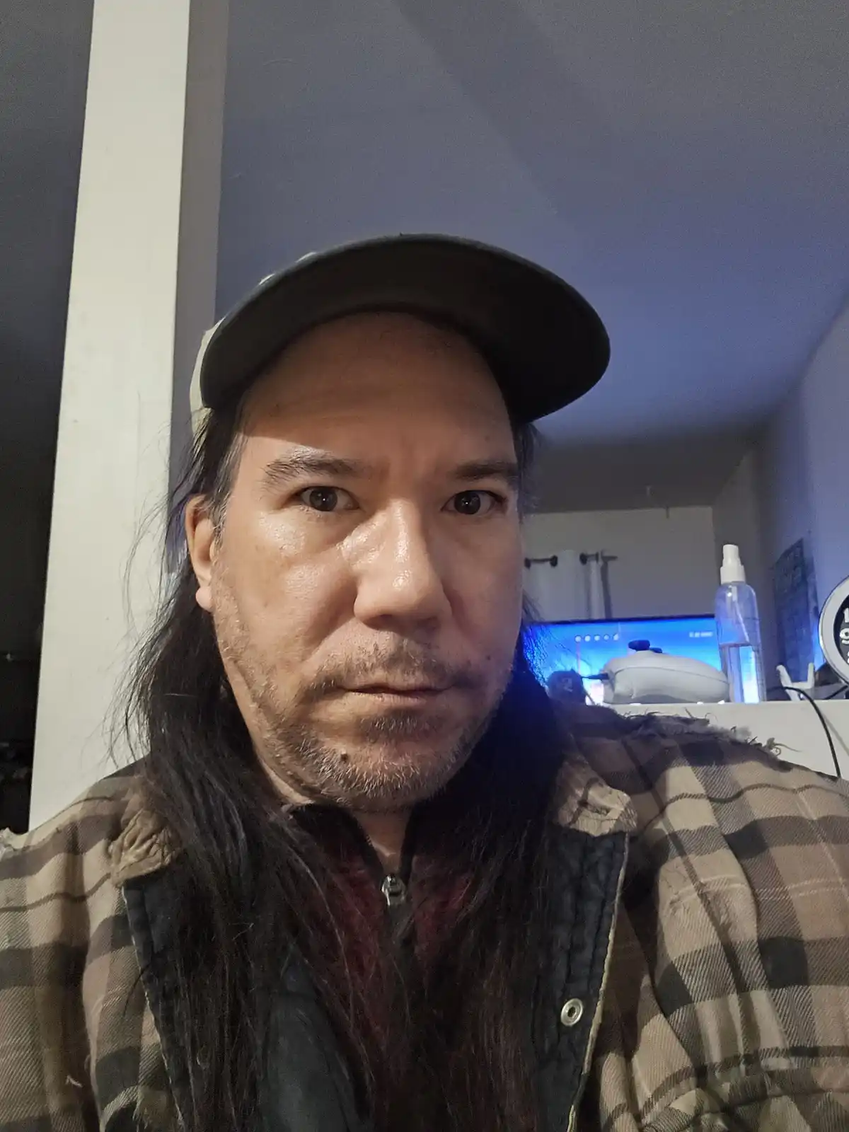 A man with long dark hair, wearing a backward cap and a plaid shirt over a vest, stares intensely at the camera with a…