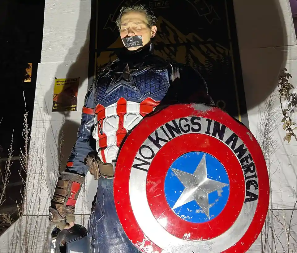 A person dressed as Captain America stands with their eyes closed and mouth covered by black tape that reads 'NO. YOU MOVE.'…