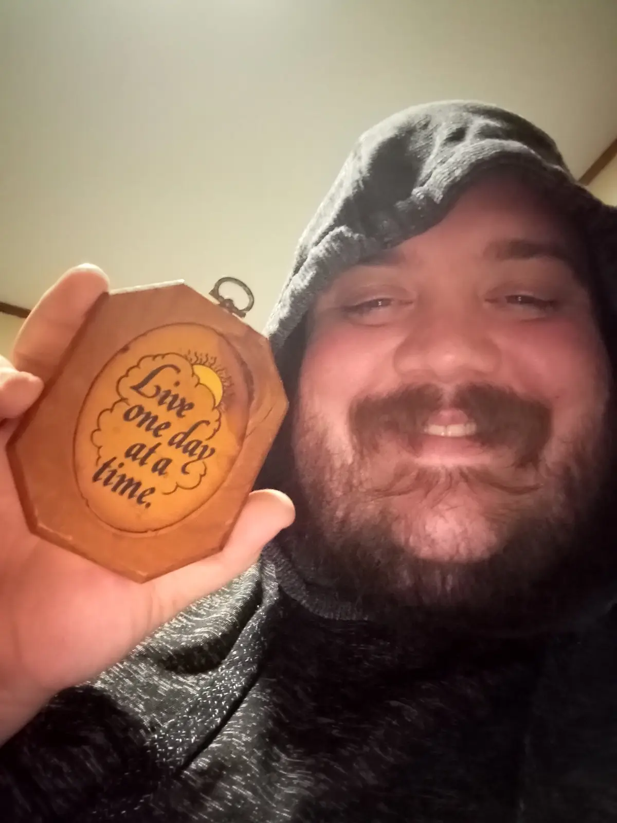 A man with a beard and hoodie smiles while holding up a wooden hexagonal plaque that reads 'Live one day at a time' with a…