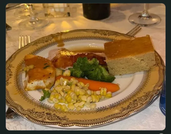 a plated meal featuring chicken, broccoli, corn, a carrot, and a slice of cornbread on an ornate gold-rimmed plate, with…