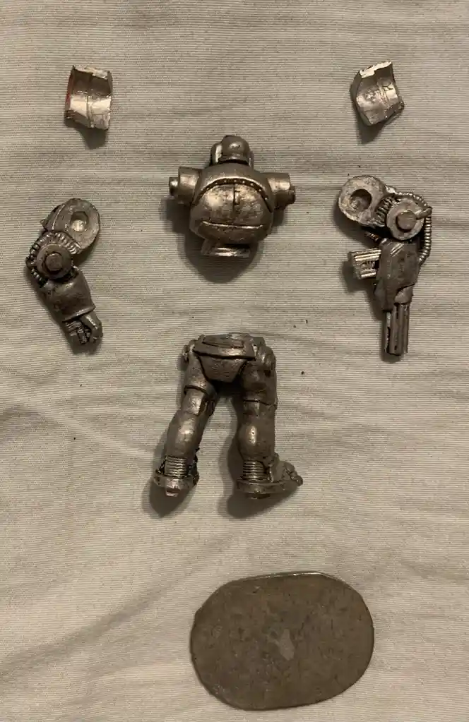 The image displays the disassembled parts of a small, silver-colored robot toy laid out on a light gray fabric surface…
