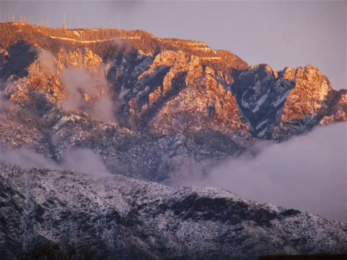 A scenic mountain landscape at sunrise or sunset, with snow-dusted peaks and clouds drifting through valleys. The sun…
