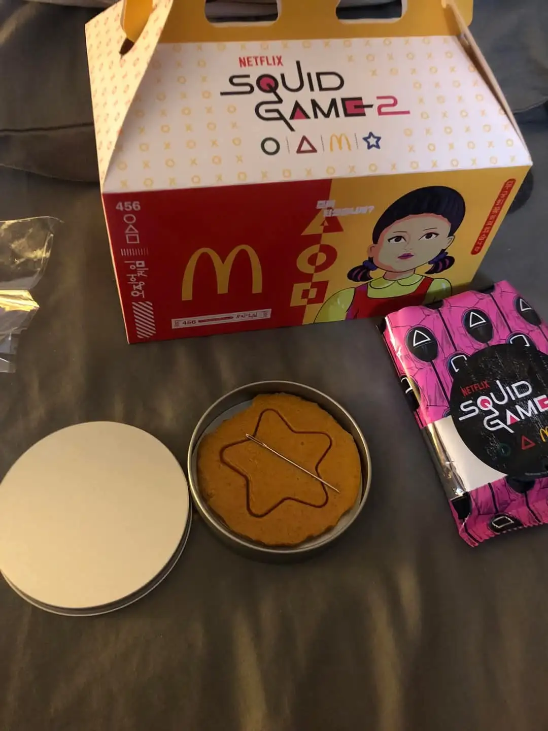 A McDonald's promotional box for 'Squid Game 2' featuring a cartoon girl and game symbols sits next to a pink snack pack and…