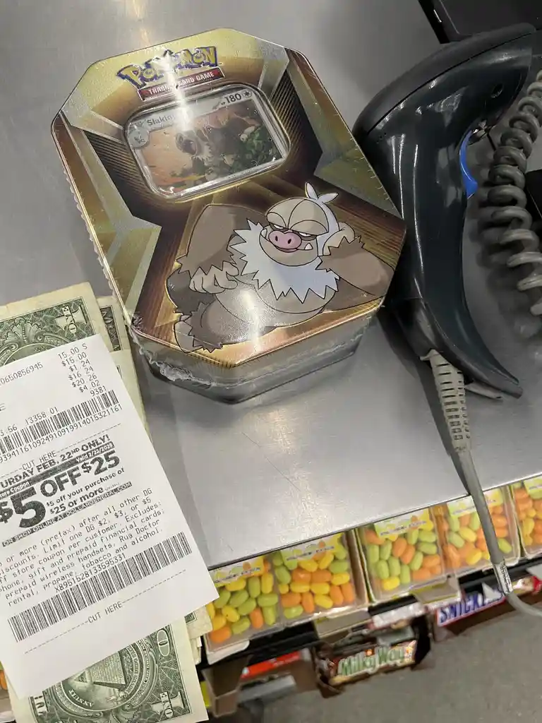 A Pokémon Trading Card Game tin featuring the Pokémon Slaking is placed on a checkout counter next to a receipt, cash, and a…