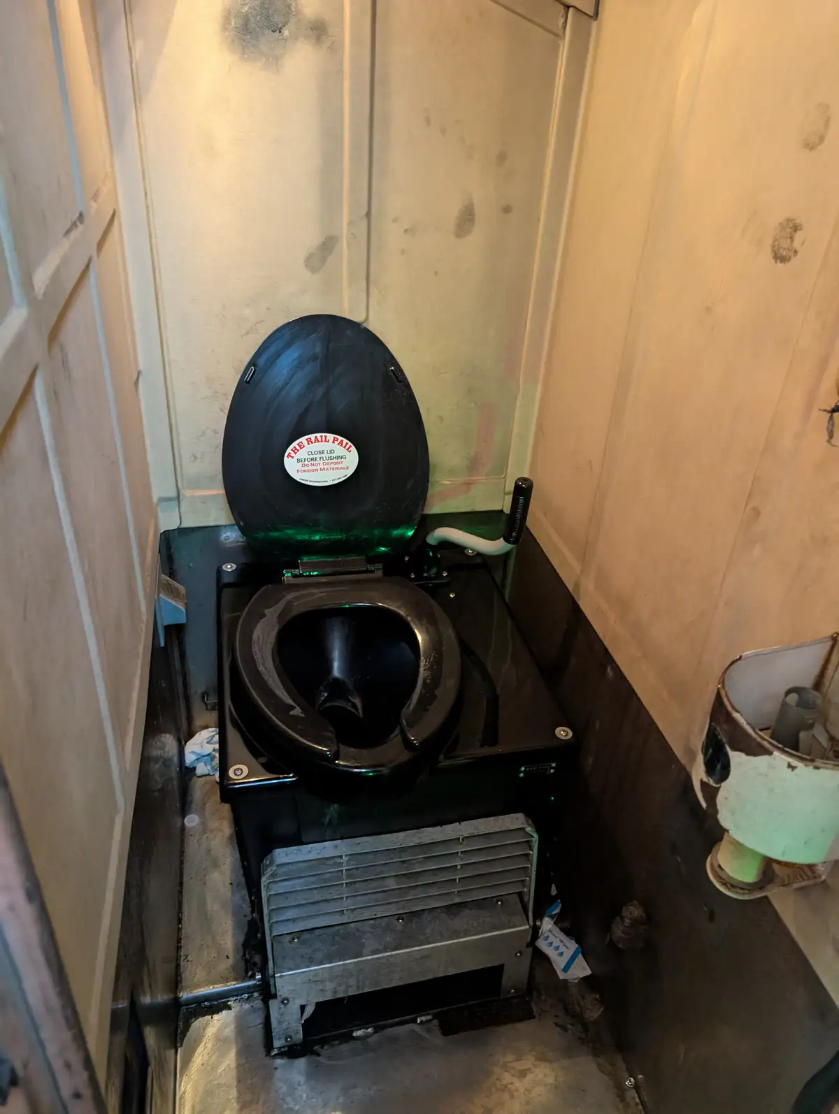 A close-up photo of a black, industrial-style toilet in a cramped, grimy stall with peeling walls. The toilet lid is open…