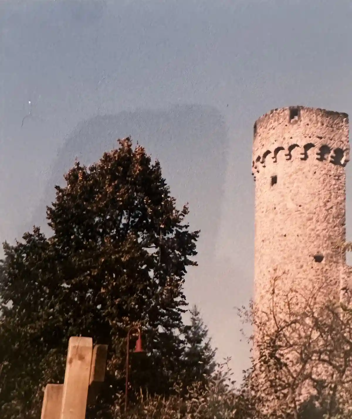 A vintage-style photo shows a tall, stone medieval tower with crenellations against a pale sky, partially framed by dark…