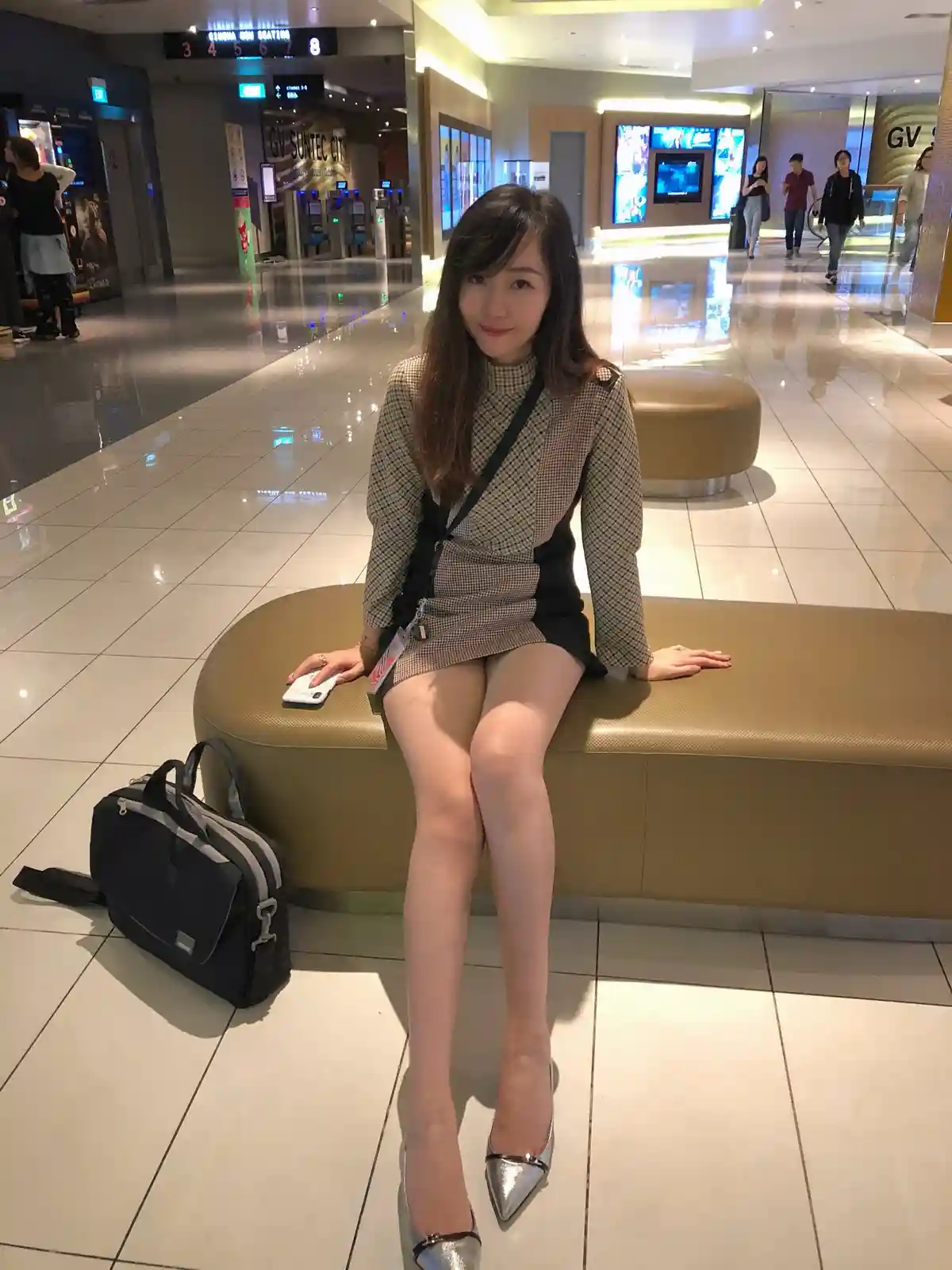 A woman sits on a curved bench in a brightly lit cinema lobby, wearing a checkered dress and silver heels, with a black bag…