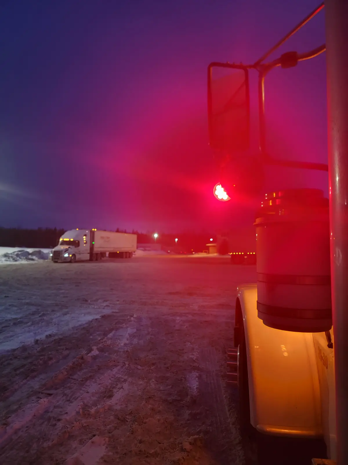 A snowy, nighttime truck stop scene viewed from the side of a truck, with a red light casting a glow over the snow-covered…