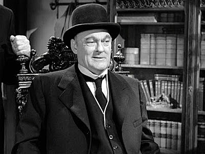 A black-and-white still of a man in a bowler hat and suit, seated in an ornate chair with bookshelves behind him, smiling…