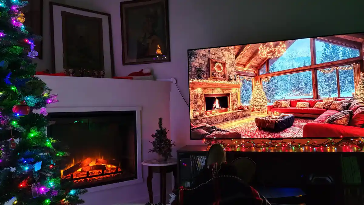 A person is sitting in a cozy, dimly lit living room decorated for Christmas with a lit tree and fireplace, watching a TV…