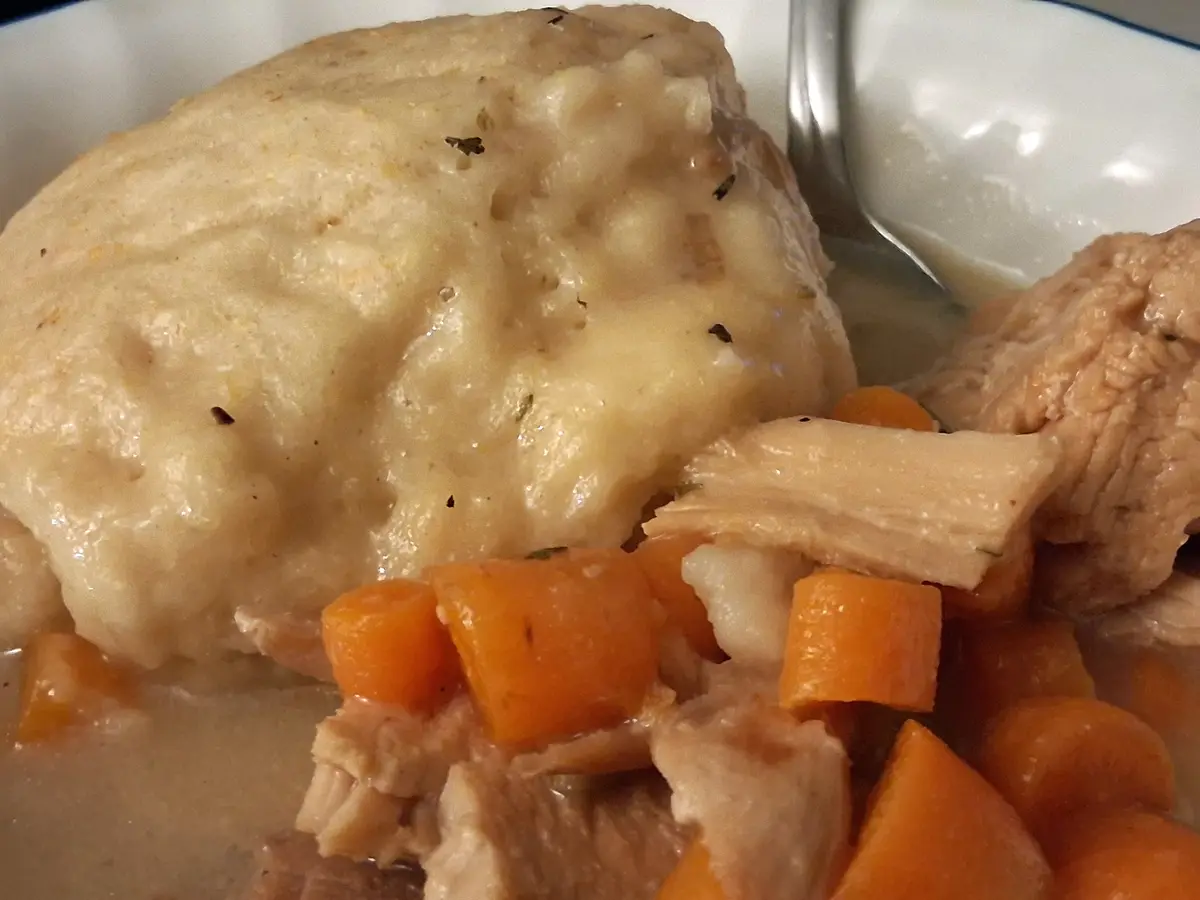 A close-up photo of a plate of food featuring a large, round, creamy mashed potato-like topping over meat and carrots, with…