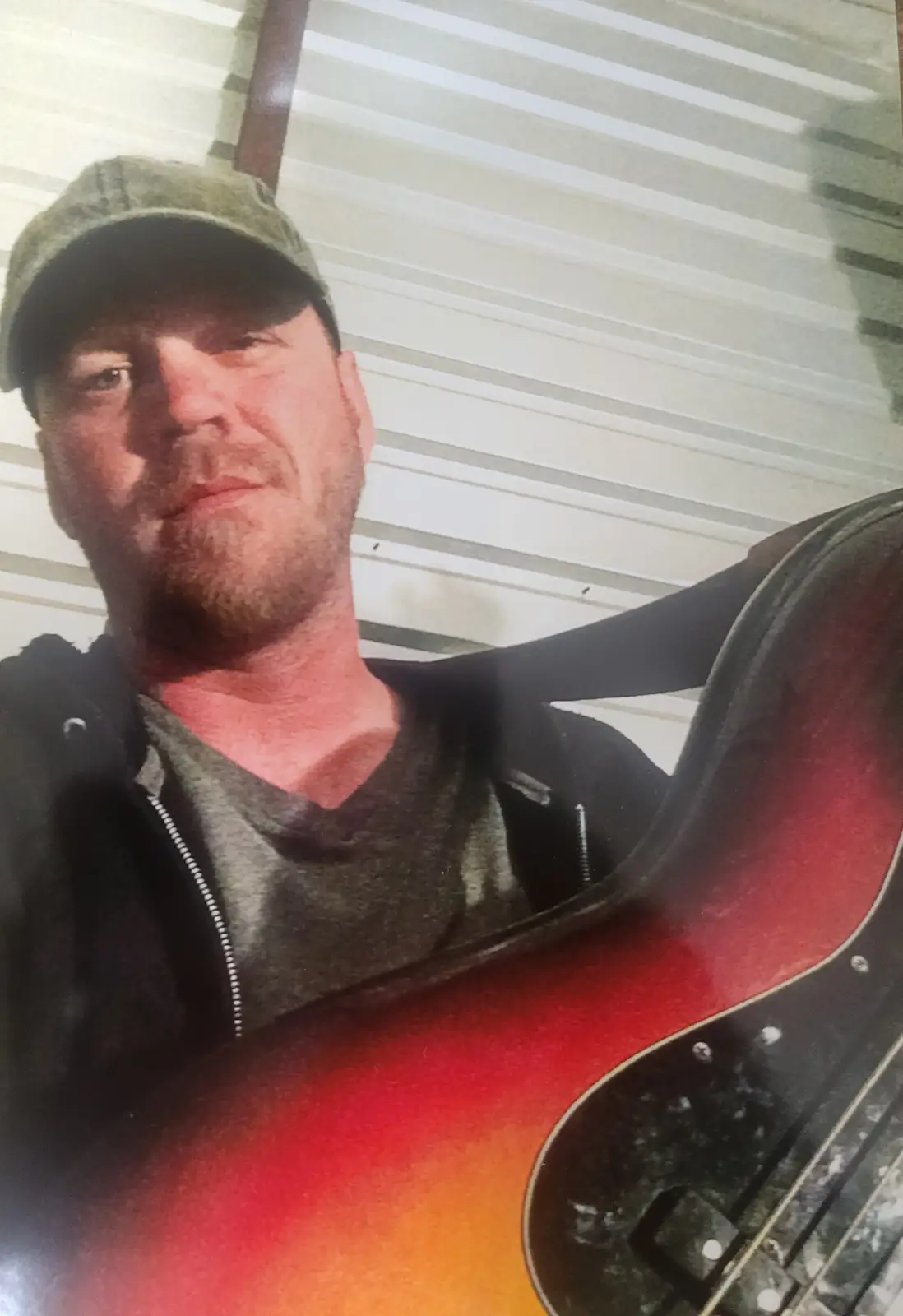 A man wearing a green cap and dark hoodie looks at the camera while holding a red and orange sunburst bass guitar. The…