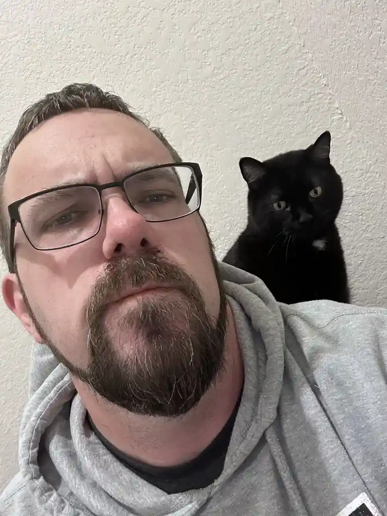 A man with glasses and a beard makes a skeptical, disappointed face while a black cat with yellow eyes sits on his shoulder…