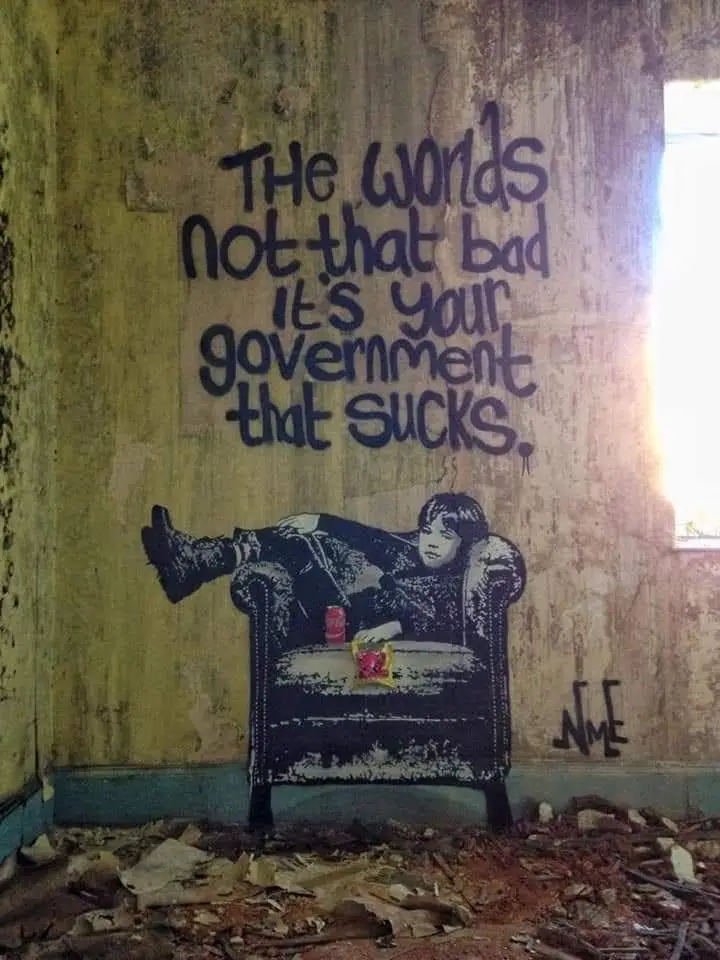 A graffiti mural on a dilapidated wall shows a boy lounging in an armchair with a soda and snack, while the text above reads…