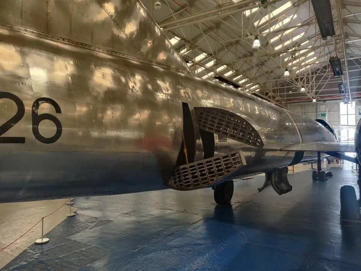A close-up photo of a silver, riveted vintage jet aircraft inside a hangar, showing its rear fuselage, engine exhaust, and…