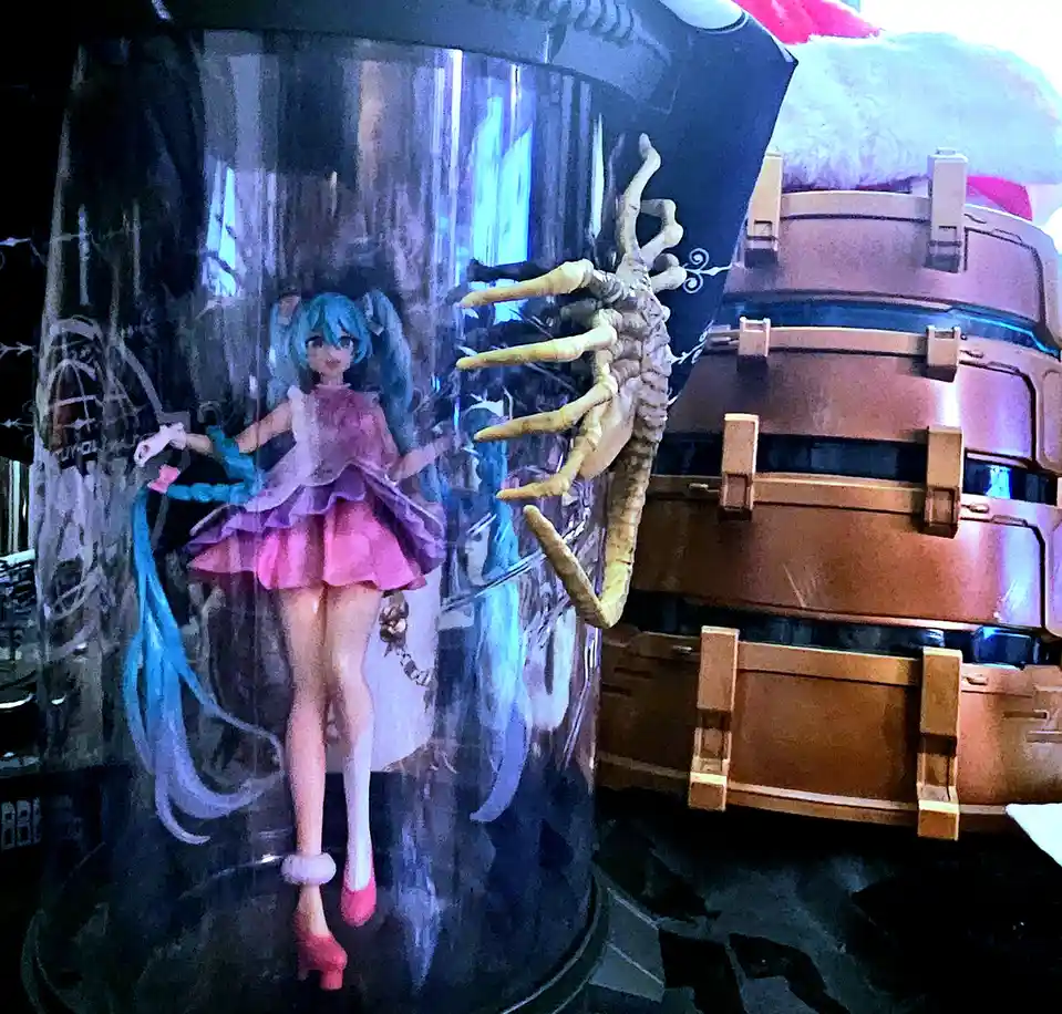 A figurine of the anime character Hatsune Miku in a pink dress stands beside a large, brown, stacked container with a…