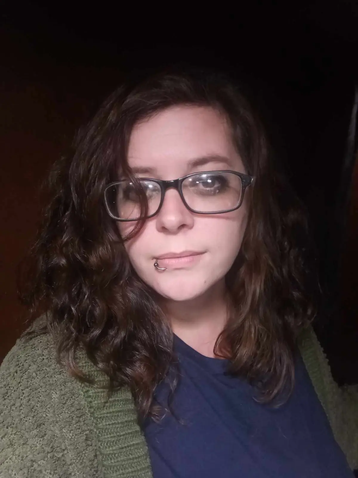 A person with dark curly hair, black-rimmed glasses, and a lip piercing stares directly at the camera with a neutral…