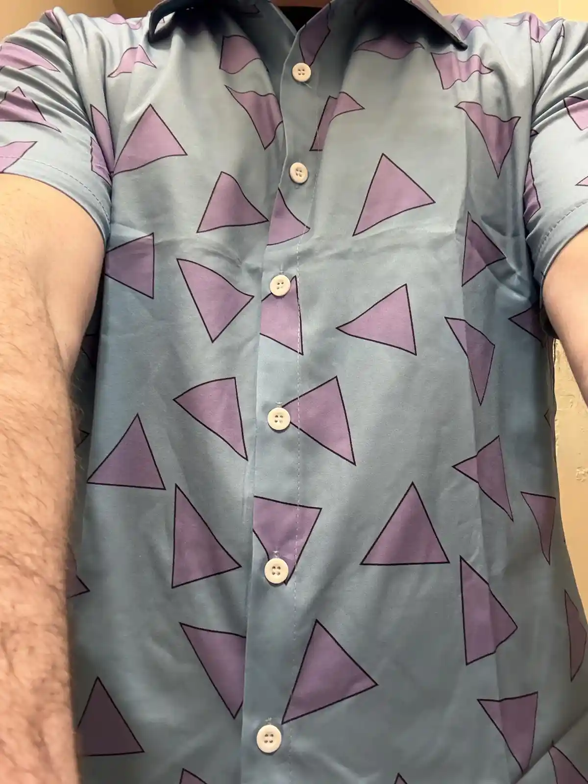 A close-up selfie shows a person wearing a light blue short-sleeved button-up shirt covered in a repeating pattern of purple…