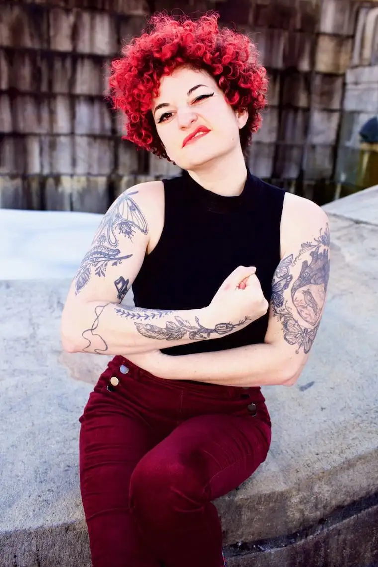 A person with bright red curly hair, heavy black eyeliner, and red lipstick sits on concrete steps wearing a black…