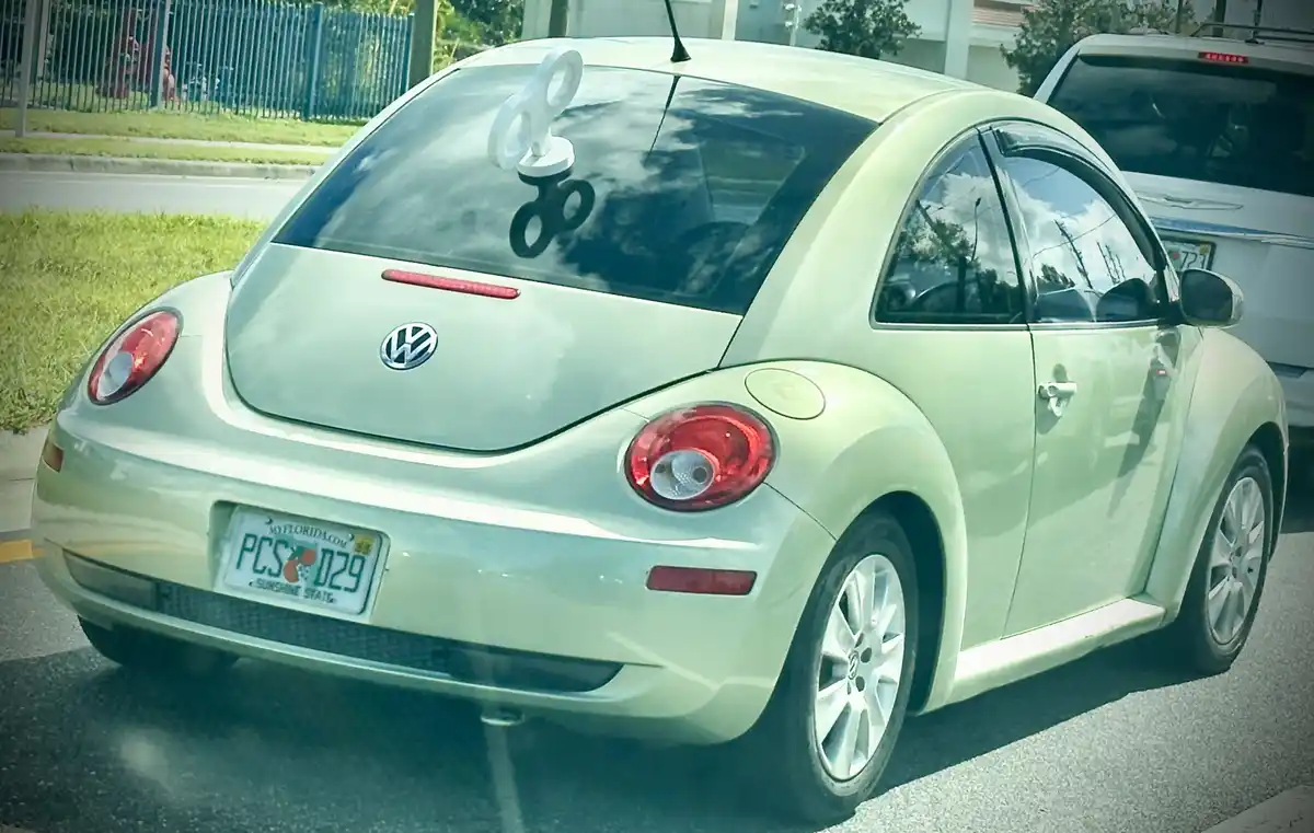 A pale green Volkswagen Beetle is parked outdoors, its rear window adorned with a white fidget spinner. The Florida license…