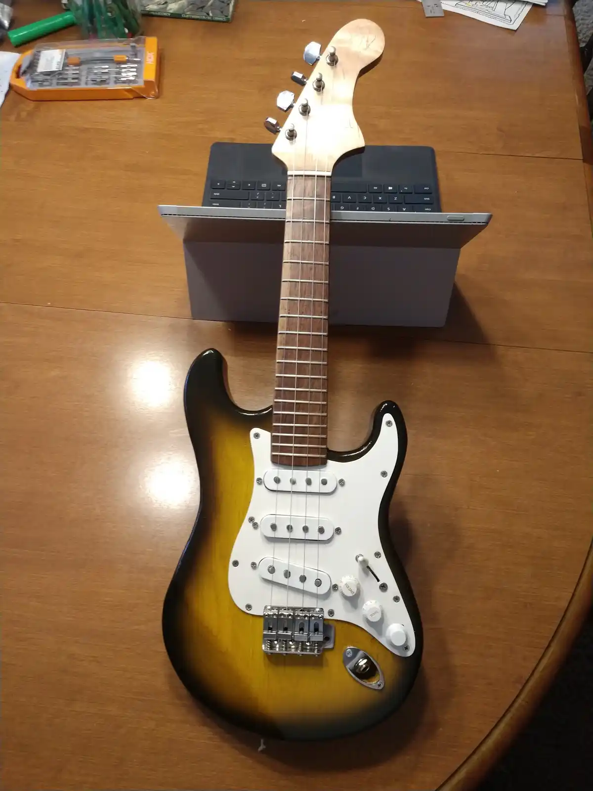 A sunburst electric guitar with a white pickguard is resting upright on a wooden table, its neck propped against the open…