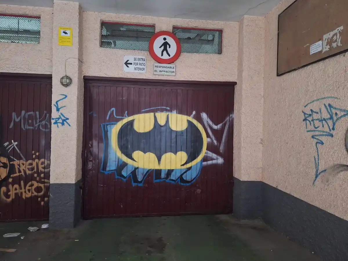 A photo of a burgundy metal gate covered in graffiti, including a large Batman logo. Above the gate, Spanish signs read 'SE…