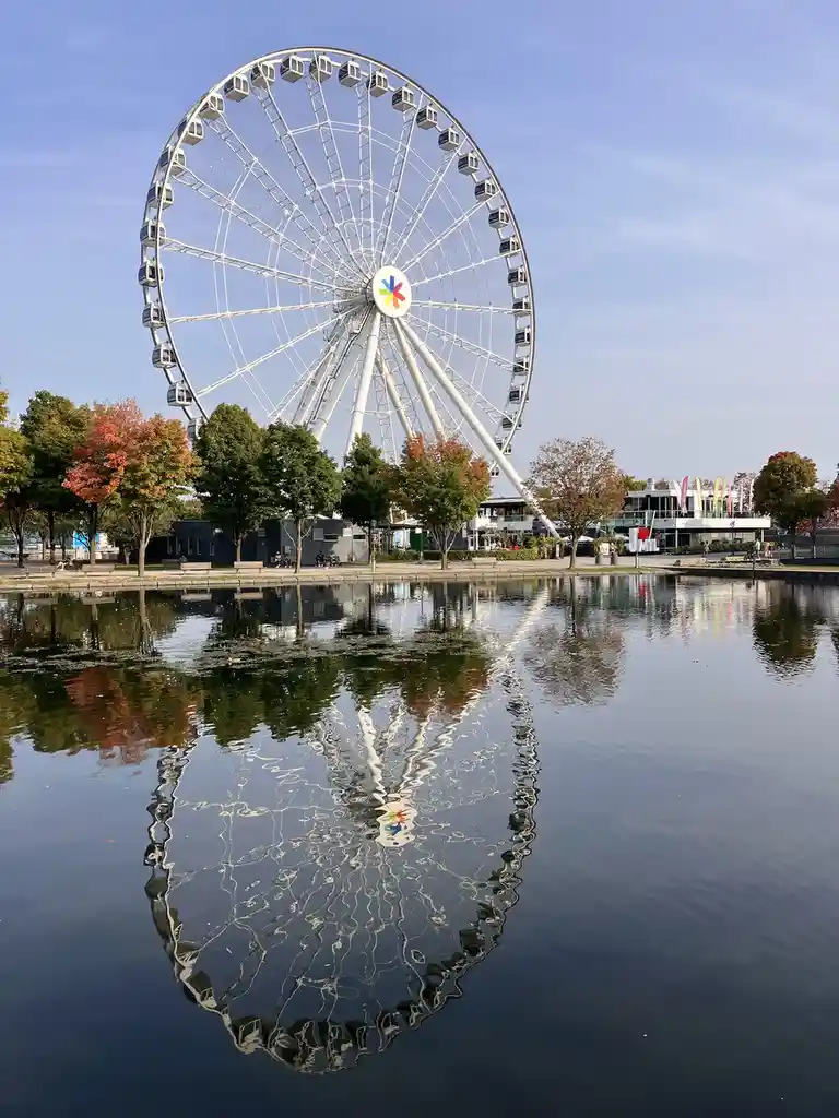 A large white Ferris wheel with a colorful logo at its center stands beside a calm body of water, its reflection distorted…