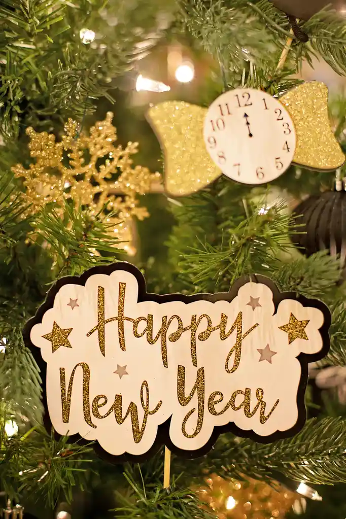 A close-up of a decorated Christmas tree with a 'Happy New Year' sign in glittery gold script on a white cloud-shaped…
